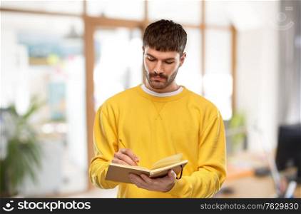 people concept - young man in yellow sweatshirt writing to diary over office background. man in yellow sweatshirt writing to diary