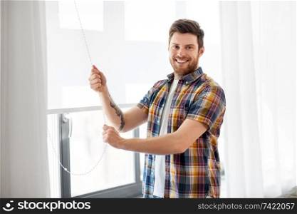 people concept - young man opening or closing roller blind on window at home. man opening roller blind on window at home