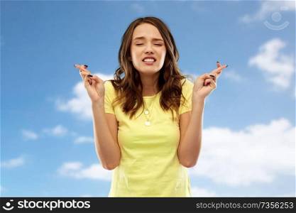 people concept - young woman or teenage girl in blank yellow t-shirt holding fingers crossed over blue sky and clouds background. young woman or teenage girl with fingers crossed
