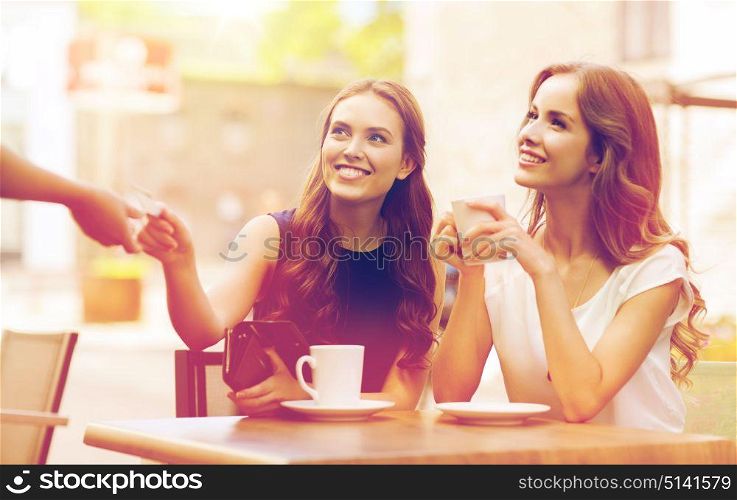 people, consumerism, lifestyle and friendship concept - smiling young women giving credit card to waiter hand and paying for coffee at outdoor cafe. women with credit card paying for coffee at cafe