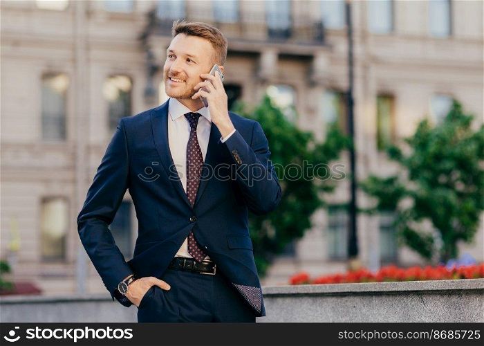 People, conversation and business concept. Handsome prosperous young male entrepreneur makes phone call in roaming, uses tariffs, stands against city blurred background, discusses something.