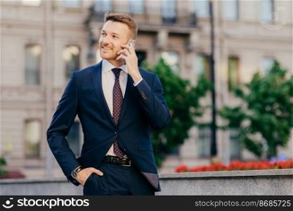 People, conversation and business concept. Handsome prosperous young male entrepreneur makes phone call in roaming, uses tariffs, stands against city blurred background, discusses something.