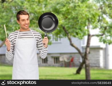 people, cooking and culinary concept - happy man or cook in apron with frying pan and wooden spoon over summer garden and house background