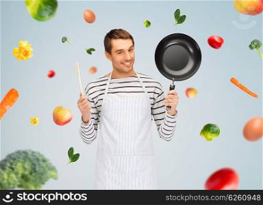 people, cooking and culinary concept - happy man or cook in apron with frying pan and wooden spoon over gray background with falling vegetables