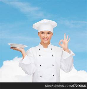 people, cooking and food concept - smiling female chef, cook or baker with empty plate showing ok sign over blue sky with cloud background