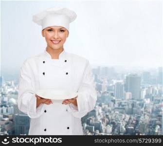 people, cooking and food concept - smiling female chef, cook or baker with empty plate over city background