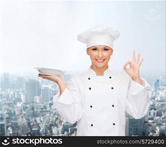 people, cooking and food concept - smiling female chef, cook or baker with empty plate showing ok sign over city background