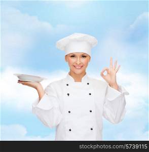 people, cooking and food concept - smiling female chef, cook or baker with empty plate showing ok sign over blue cloudy sky background