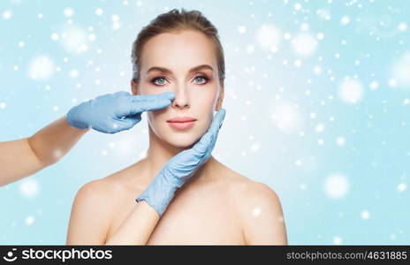 people, cosmetology, plastic surgery and beauty concept - surgeon or beautician hands touching woman face over blue background and snow