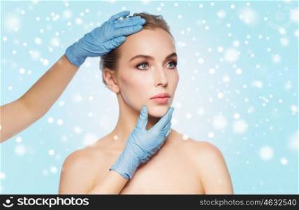 people, cosmetology, plastic surgery and beauty concept - surgeon or beautician hands touching woman face over blue background and snow