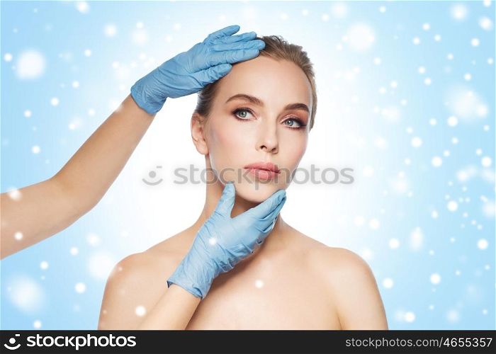 people, cosmetology, plastic surgery and beauty concept - surgeon or beautician hands touching woman face over blue background and snow