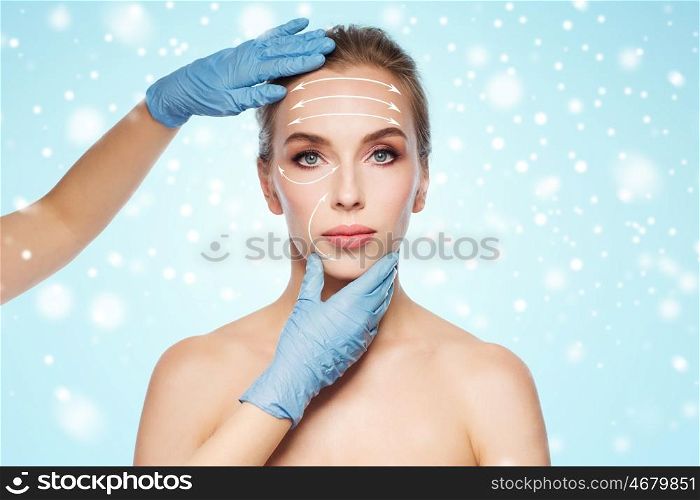 people, cosmetology, plastic surgery and beauty concept - surgeon or beautician hands touching woman face with lifting arrows over blue background and snow