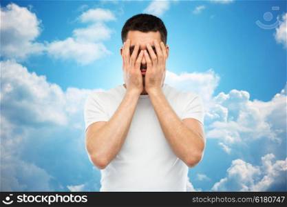 people, crisis, emotions and stress concept - man in white t-shirt covering his face with hands over blue sky and clouds background