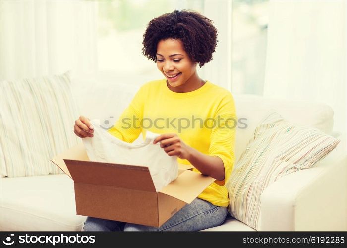people, delivery, commerce, shipping and postal service concept - happy african american young woman taking clothes out of cardboard box or parcel at home