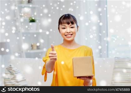 people, delivery, shipping, gesture and shopping concept - happy asian young woman with cardboard parcel box showing thumbs up at home over snow