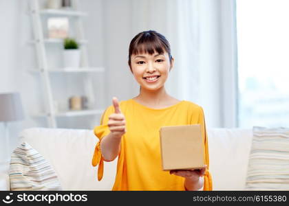 people, delivery, shipping, gesture and shopping concept - happy asian young woman with cardboard parcel box showing thumbs up at home