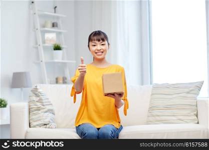 people, delivery, shipping, gesture and shopping concept - happy asian young woman with cardboard parcel box showing thumbs up at home