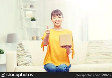 people, delivery, shipping, gesture and shopping concept - happy asian young woman with cardboard parcel box showing thumbs up at home. happy asian young woman with parcel box at home. happy asian young woman with parcel box at home