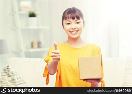 people, delivery, shipping, gesture and shopping concept - happy asian young woman with cardboard parcel box showing thumbs up at home. happy asian young woman with parcel box at home