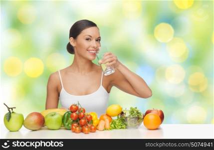 people, diet, healthy eating and food concept - happy woman drinking water from glass with fruits and vegetables on table over green summer lights background. happy woman with glass of water and healthy food