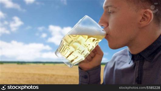 people, drinks, alcohol and leisure concept - close up of young man drinking beer from glass mug over cereal field and blue sky background. close up of young man drinking beer from glass mug
