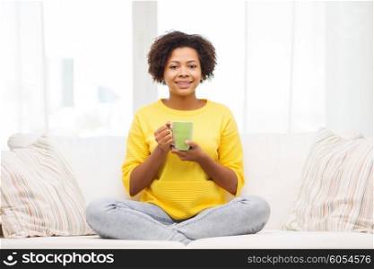 people, drinks and leisure concept - happy african american woman sitting on sofa and drinking tea from cup or mug at home