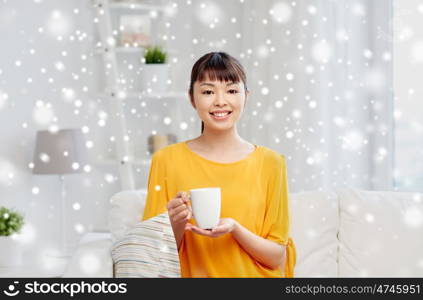 people, drinks and leisure concept - happy asian woman sitting on sofa and drinking tea from cup or mug at home over snow