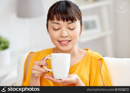 people, drinks and leisure concept - happy asian woman sitting on sofa and drinking tea from cup or mug at home