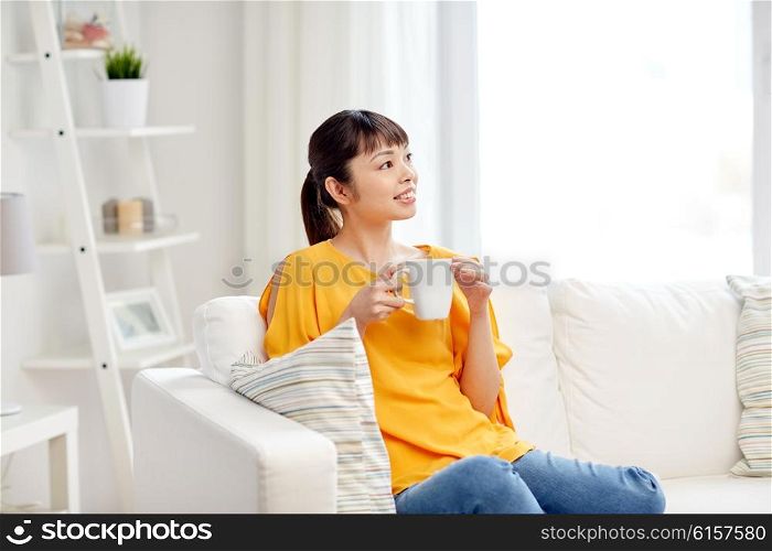 people, drinks and leisure concept - happy asian woman sitting on sofa and drinking tea from cup or mug at home