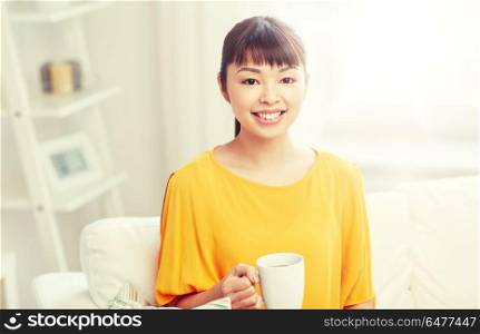 people, drinks and leisure concept - happy asian woman sitting on sofa and drinking tea from cup or mug at home. happy asian woman drinking from tea cup. happy asian woman drinking from tea cup