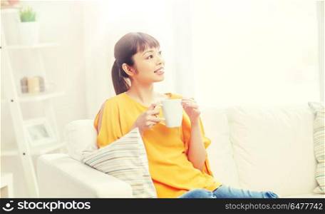 people, drinks and leisure concept - happy asian woman sitting on sofa and drinking tea from cup or mug at home. happy asian woman drinking from tea cup. happy asian woman drinking from tea cup