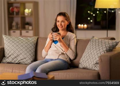 people, drinks and leisure concept - happy woman with mug drinking tea or coffee at home in evening. happy woman drinking tea or coffee at home