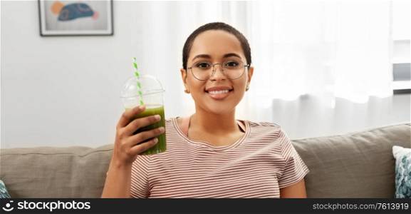 people, eating and leisure concept - happy smiling african american young woman sitting on sofa and drinking smoothie from plastic cup with paper straw at home. african american woman drinking smoothie at home