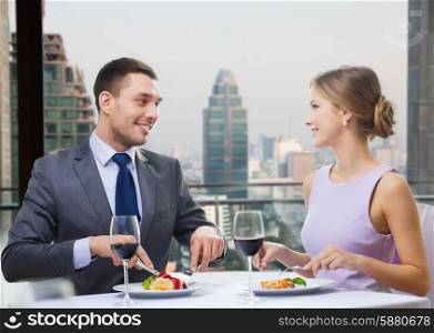 people, eating, celebration, romantic and holidays concept - smiling couple with red wine and food talking at restaurant over city background