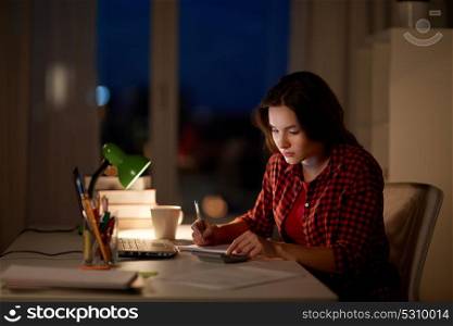 people, education, freelance and learning concept - student girl with notebook and calculator doing math at home at night. student girl with notebook and calculator at home