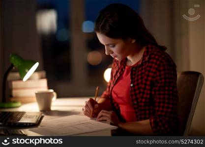 people, education, freelance and learning concept - woman or student girl with laptop computer writing to notebook at home at night. student girl with laptop and notebook at home
