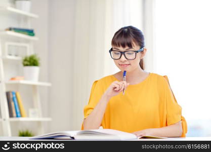 people, education, high school and learning concept - happy asian young woman student in glasses reading book and writing to notepad at home
