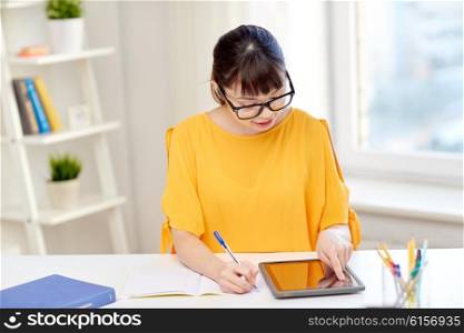 people, education, high school and learning concept - happy asian young woman student in glasses with tablet pc computer, book and notepad writing at home