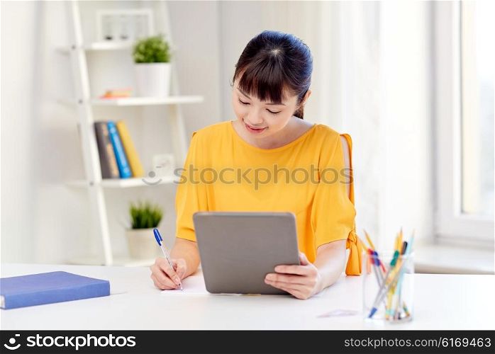 people, education, high school and learning concept - happy asian young woman student with tablet pc computer, book and notepad writing at home