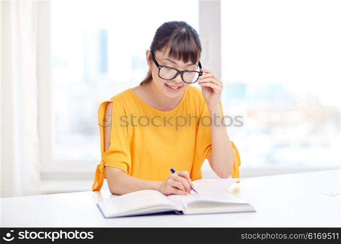 people, education, high school and learning concept - happy asian young woman student in glasses reading book and writing to notepad at home
