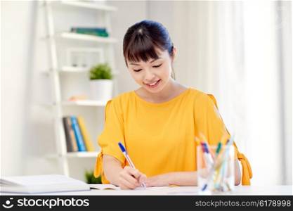 people, education, high school and learning concept - happy asian young woman student with book and notepad writing at home