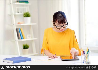 people, education, high school and learning concept - happy asian young woman student in glasses with tablet pc computer, book and notepad writing at home