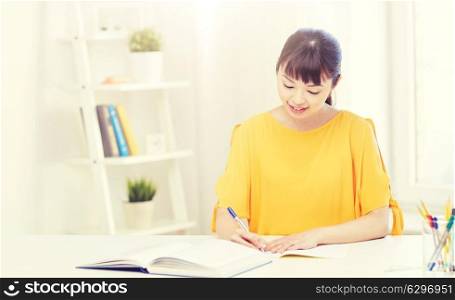 people, education, high school and learning concept - happy asian young woman student with book and notepad writing at home. happy asian young woman student learning at home. happy asian young woman student learning at home