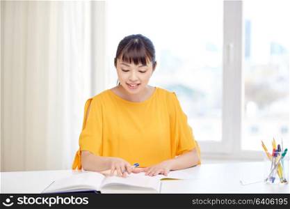 people, education, high school and learning concept - happy asian young woman student with book and notepad writing at home. happy asian young woman student learning at home