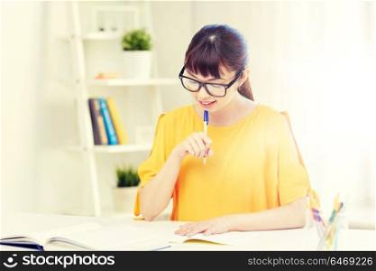 people, education, high school and learning concept - happy asian young woman student in glasses reading book and writing to notepad at home. happy asian young woman student learning at home