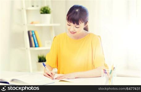 people, education, high school and learning concept - happy asian young woman student with book and notepad writing at home. happy asian young woman student learning at home. happy asian young woman student learning at home
