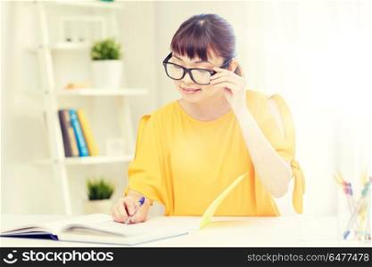 people, education, high school and learning concept - happy asian young woman student in glasses reading book and writing to notepad at home. happy asian young woman student learning at home. happy asian young woman student learning at home