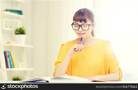 people, education, high school and learning concept - happy asian young woman student in glasses reading book and writing to notepad at home. happy asian young woman student learning at home. happy asian young woman student learning at home