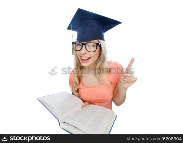 people, education, knowledge and graduation concept - smiling young student woman in mortarboard and eyeglasses with encyclopedia book pointing finger up