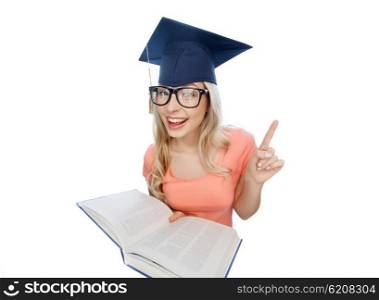 people, education, knowledge and graduation concept - smiling young student woman in mortarboard and eyeglasses with encyclopedia book pointing finger up
